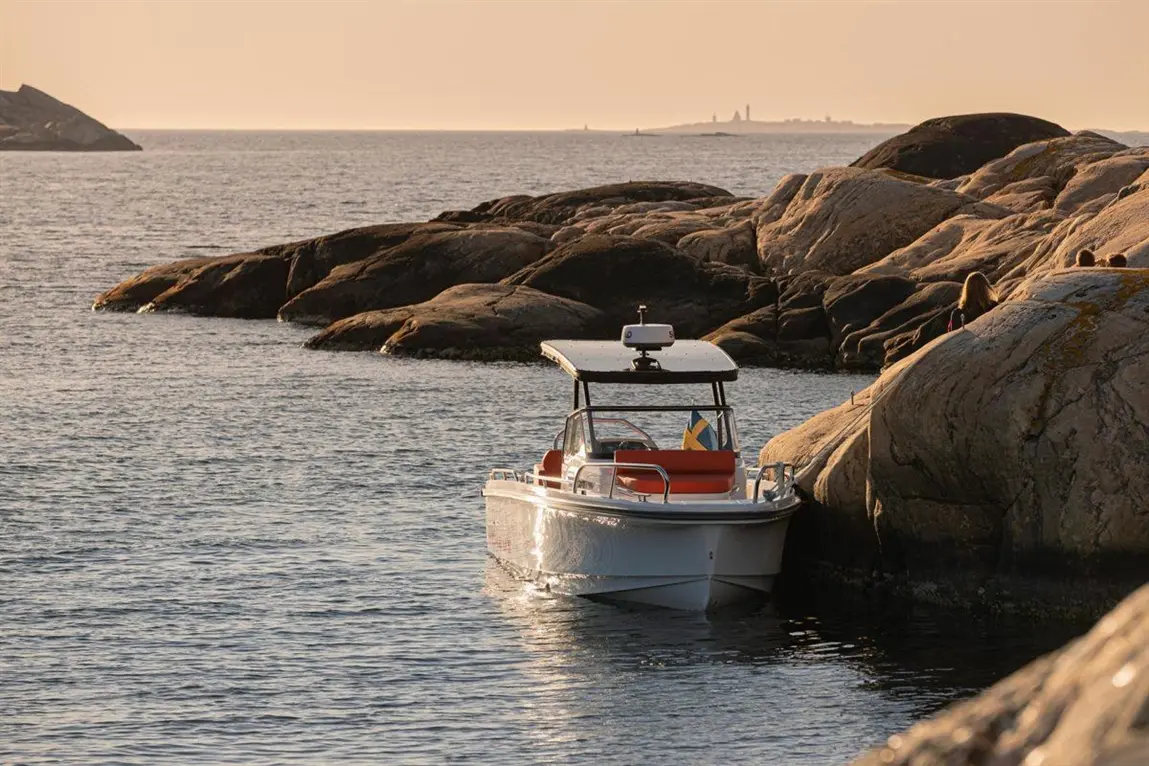 A small boat with a Swedish flag rests near rocky shores at sunset, reflecting warm colours on calm waters.